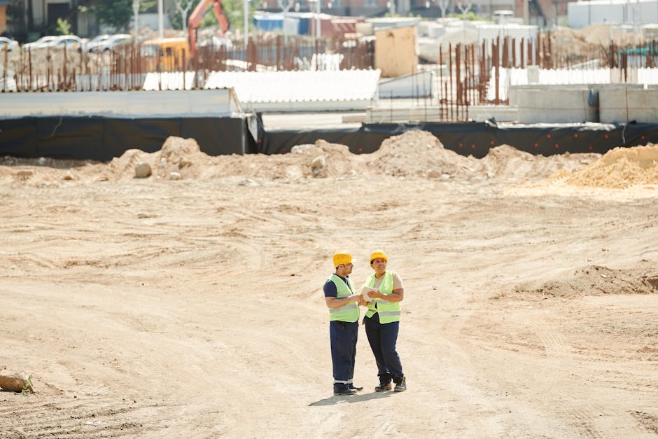 Two workers discussing plans on a sandy construction site, wearing safety gear